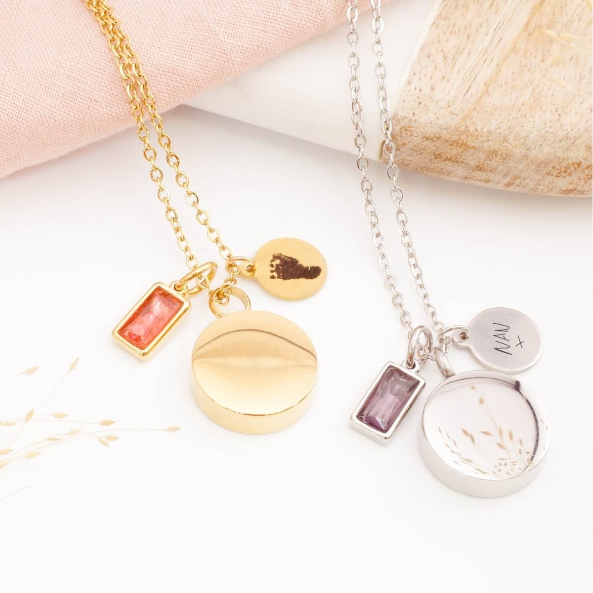 Two self fill urn necklaces with engraved footprint, handwriting tags and birthstone charms on a white surface