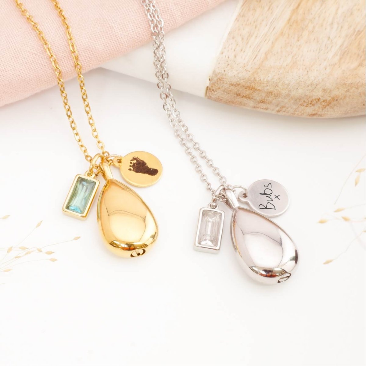 Two teardrop urn necklaces with engraved footprint, handwriting and birthstone charms on a white surface