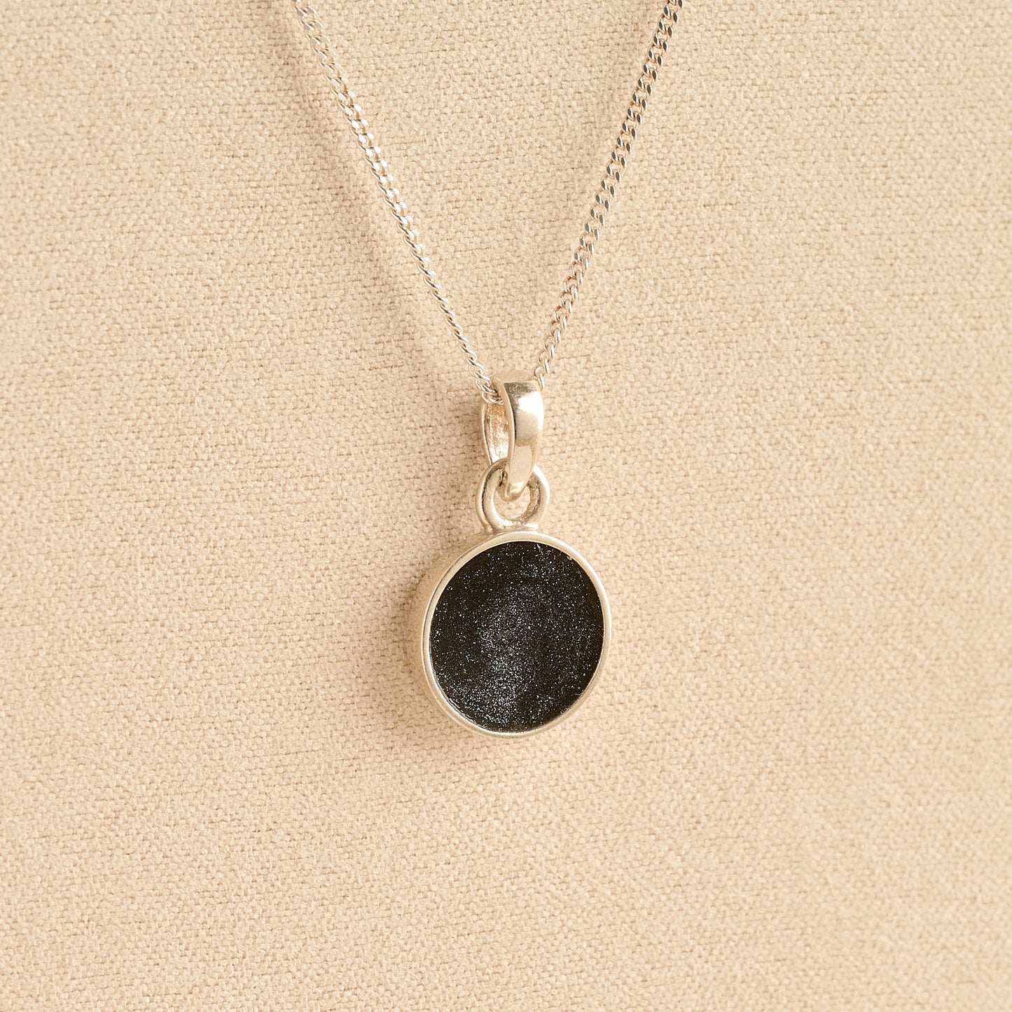 Silver Cremation ashes necklace with a round pendant and black resin filled with ashes or fur, displayed on a soft beige background – suitable for pet or human memorials.