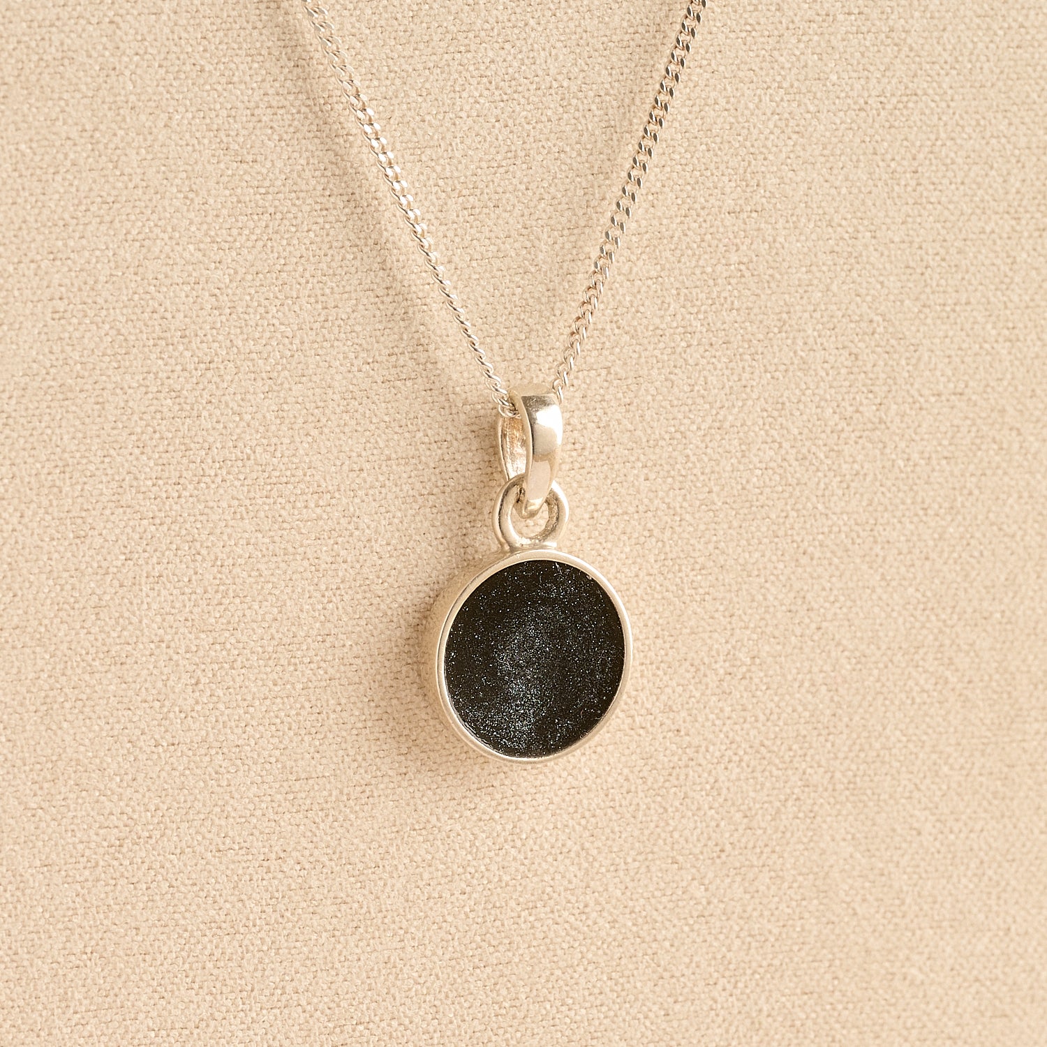 Silver Cremation ashes necklace with a round pendant and black resin filled with ashes or fur, displayed on a soft beige background – suitable for pet or human memorials.