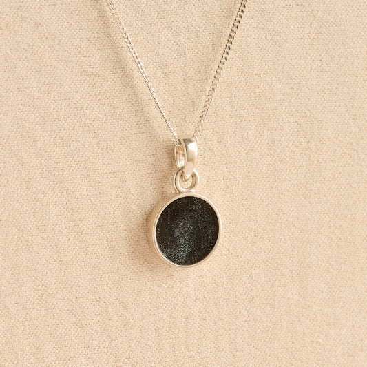 Silver Cremation ashes necklace with a round pendant and black resin filled with ashes or fur, displayed on a soft beige background – suitable for pet or human memorials.