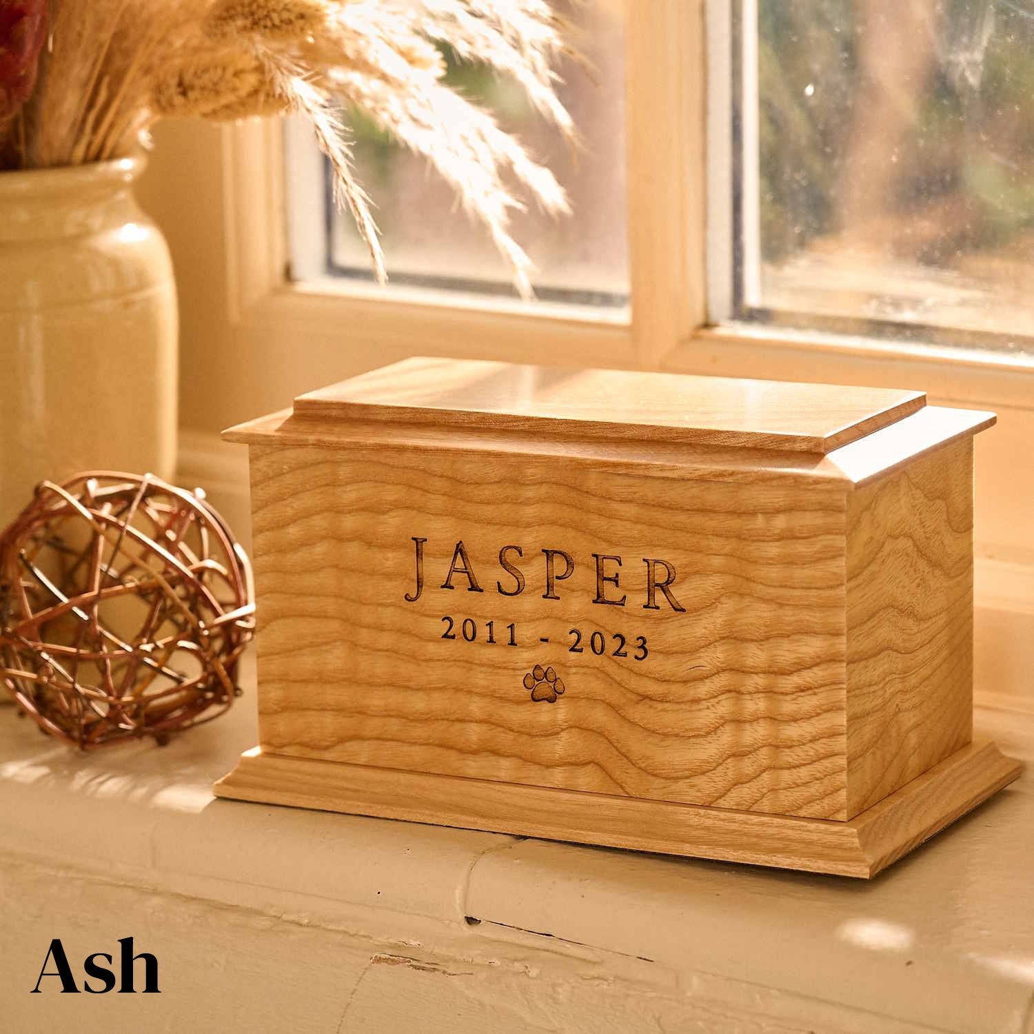Pet cremation urn made from ash wood with custom engraving. The cremation urn is wooden and sitting on a windowsill.  It has the name of the pet engraved on the front face and a paw print. It is a dog urn or a cat urn.