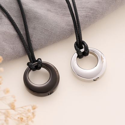 Two necklaces with circular memorial pendants on black cords, one black and one silver, on a textured surface.