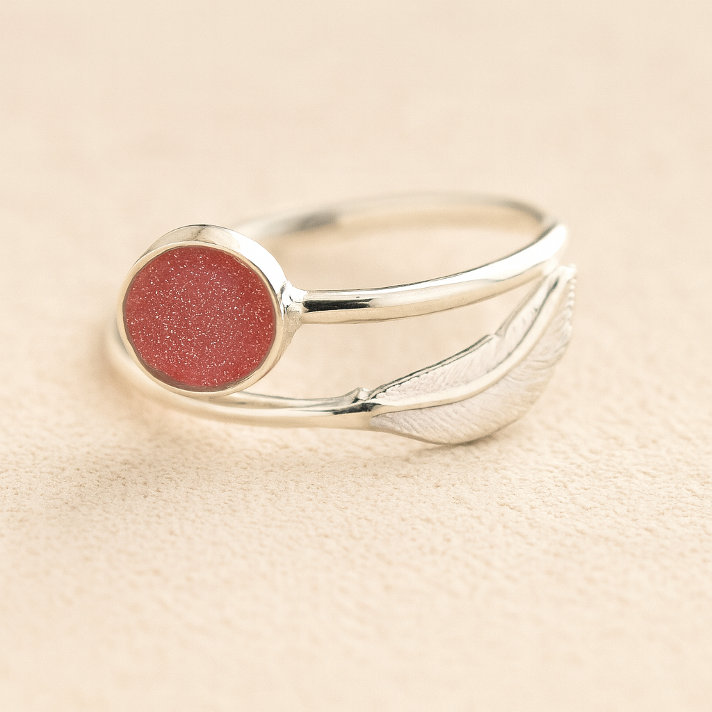 Adjustable Silver cremation ring with a dark pink resin and feather on a beige background