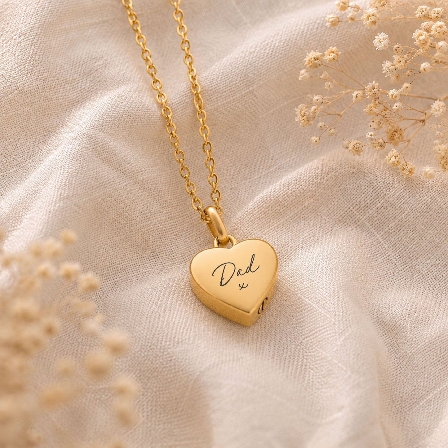Gold heart-shaped cremation necklace with 'Dad' engraving on a beige fabric background