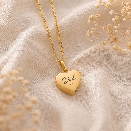 Gold heart-shaped cremation necklace with 'Dad' engraving on a beige fabric background
