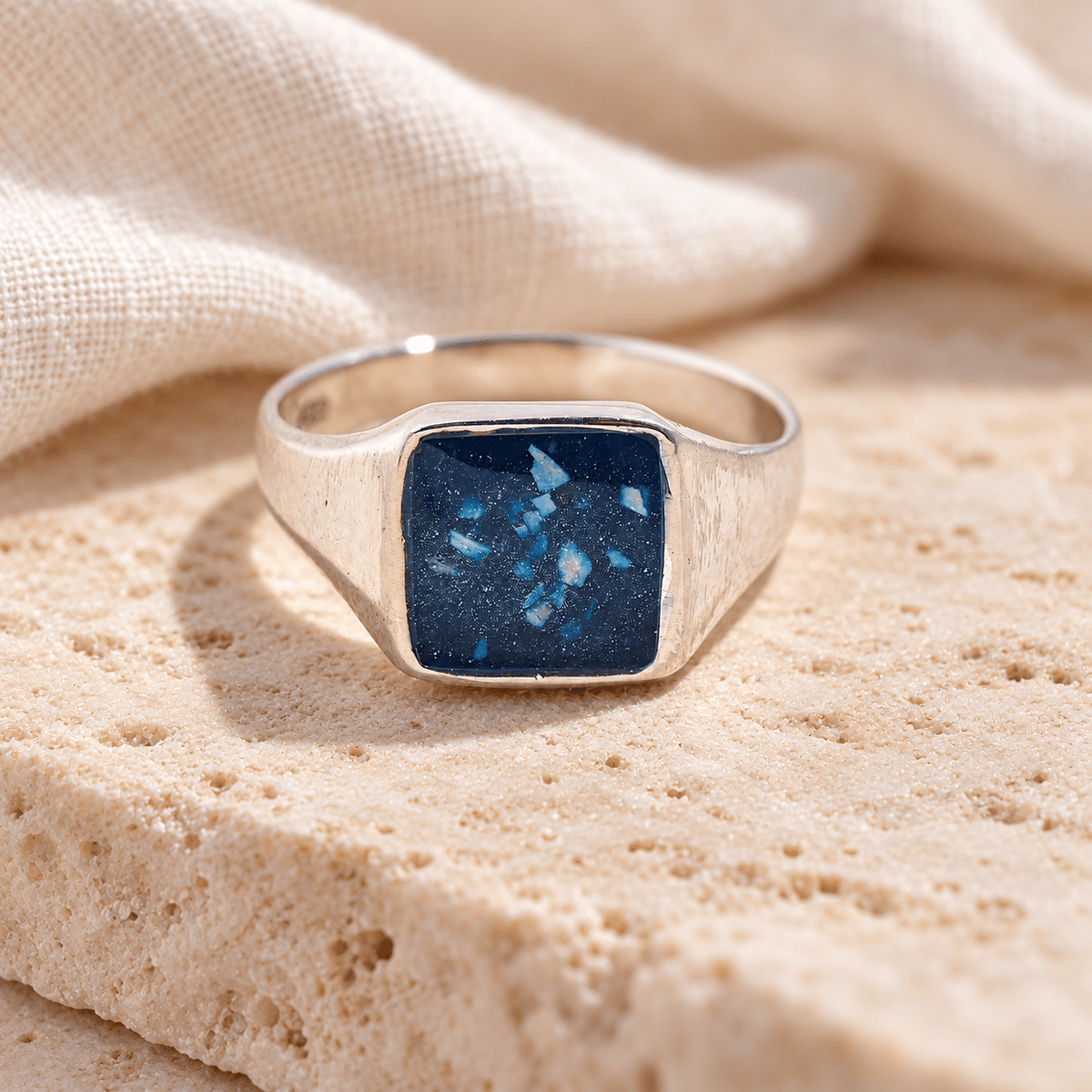 Men’s sterling silver signet ashes ring with broad tapered shoulders and a rectangular deep blue resin setting containing visible ashes, resting on cream stone.