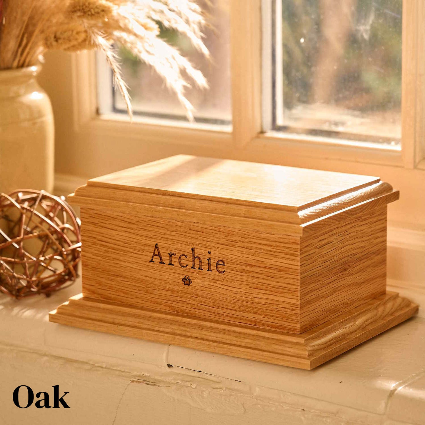 Personalised pet cremation urn crafted from oak wood. The oak urn sits on a windowsill. It has the name Archie and a pawprint engraved on it.