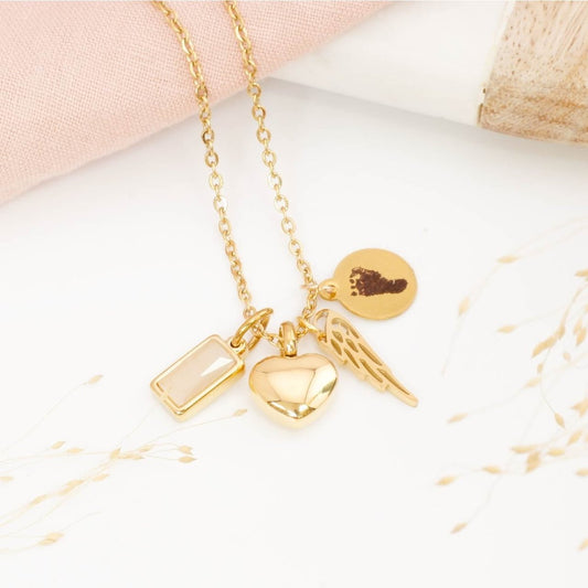 Gold footprint necklace, angel wing and heart urn pendant and birthstone charm on a light background