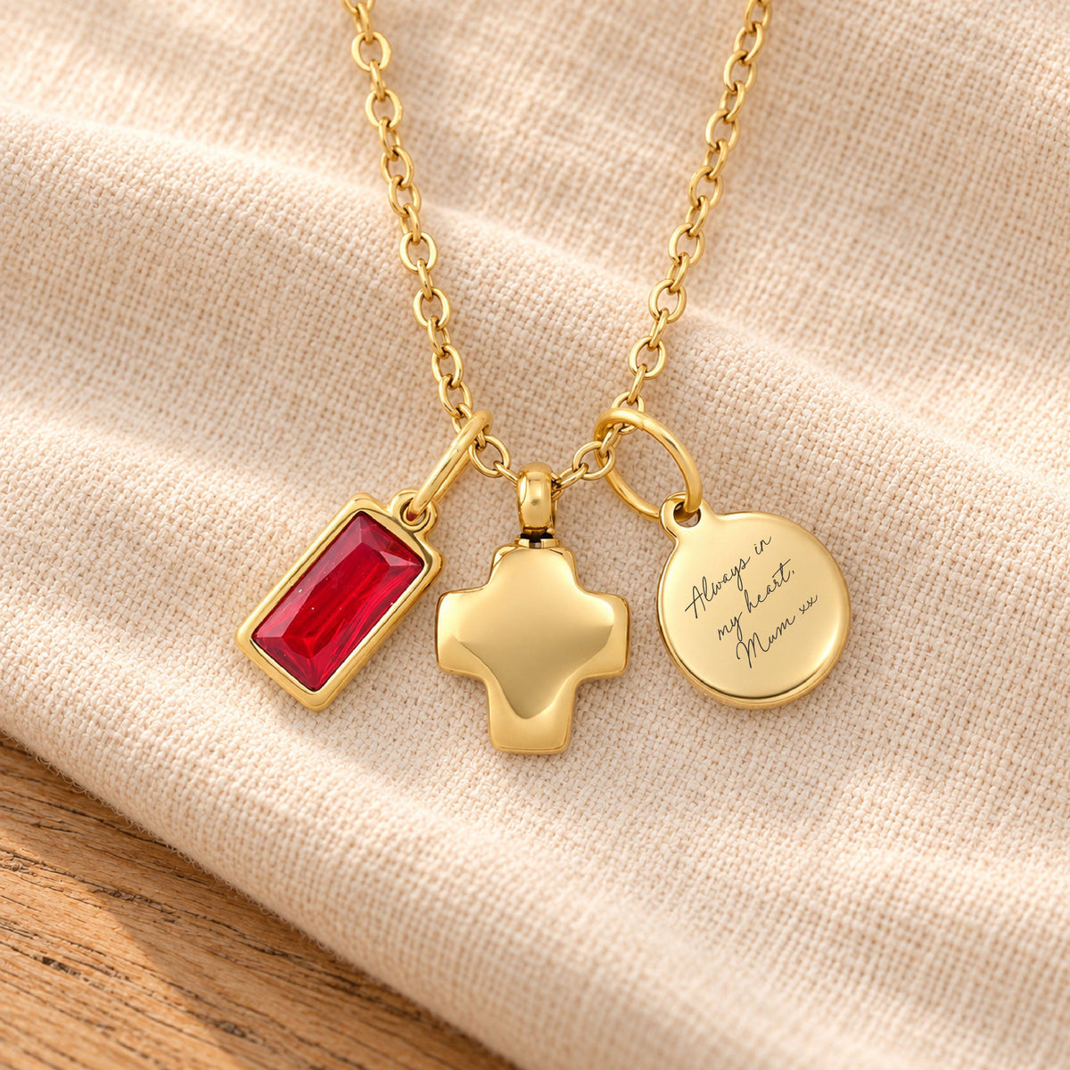 Gold memorial necklace with three charms: a rectangular red gemstone pendant, a gold cross pendant, and a round engraved disc reading 'Always in my heart Mom'. The necklace is displayed on a textured beige fabric with a wooden surface visible in the corner.