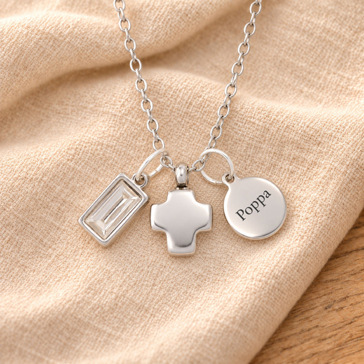 Silver-plated necklace with a rectangular clear birthstone charm, a softly rounded cross urn pendant and a blank round tag, resting on beige linen over rustic wood.
