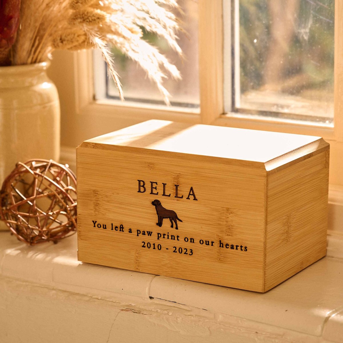 Bamboo wood dog urn with Labrador engraving and the name Bella – personalised pet cremation urn on a window sill