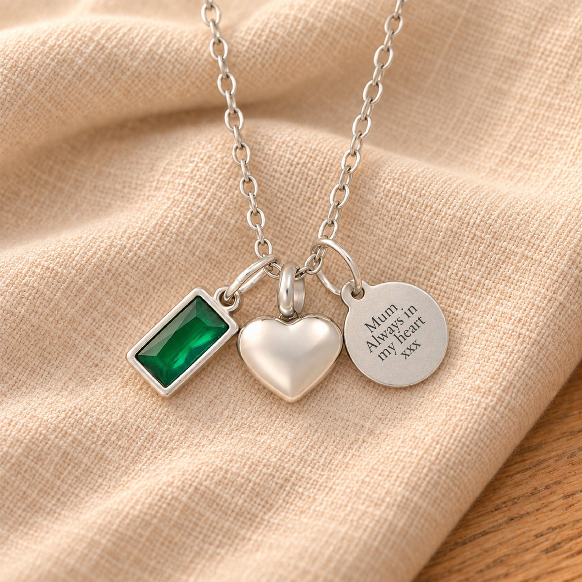 Silver-plated necklace with a rectangular green birthstone charm, a rounded heart urn pendant and a blank round tag, resting on draped beige linen.