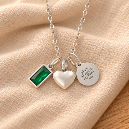 Silver-plated necklace with a rectangular green birthstone charm, a rounded heart urn pendant and a blank round tag, resting on draped beige linen.