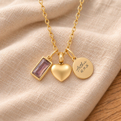 Gold-plated necklace with a rectangular pale purple birthstone charm, a rounded heart urn pendant and a blank round tag, resting on draped beige linen.
