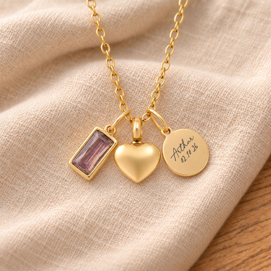 Gold-plated necklace with a rectangular pale purple birthstone charm, a rounded heart urn pendant and a blank round tag, resting on draped beige linen.