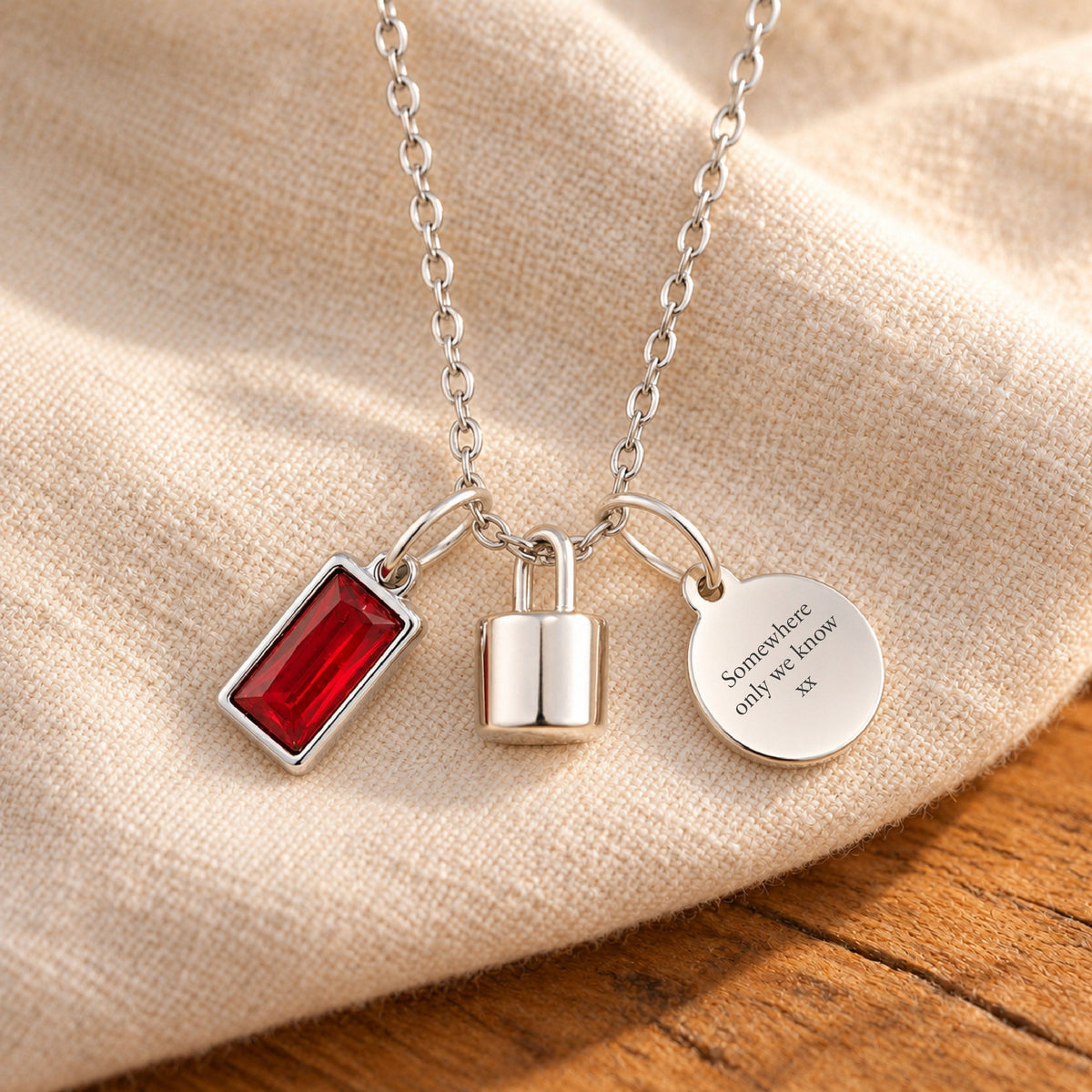 Silver-plated necklace with a rectangular red birthstone charm, a polished lock-shaped urn pendant and a blank round tag, resting on beige linen over rustic wood.