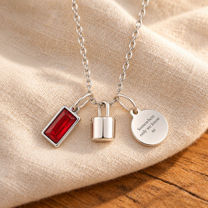 Silver-plated necklace with a rectangular red birthstone charm, a polished lock-shaped urn pendant and a blank round tag, resting on beige linen over rustic wood.