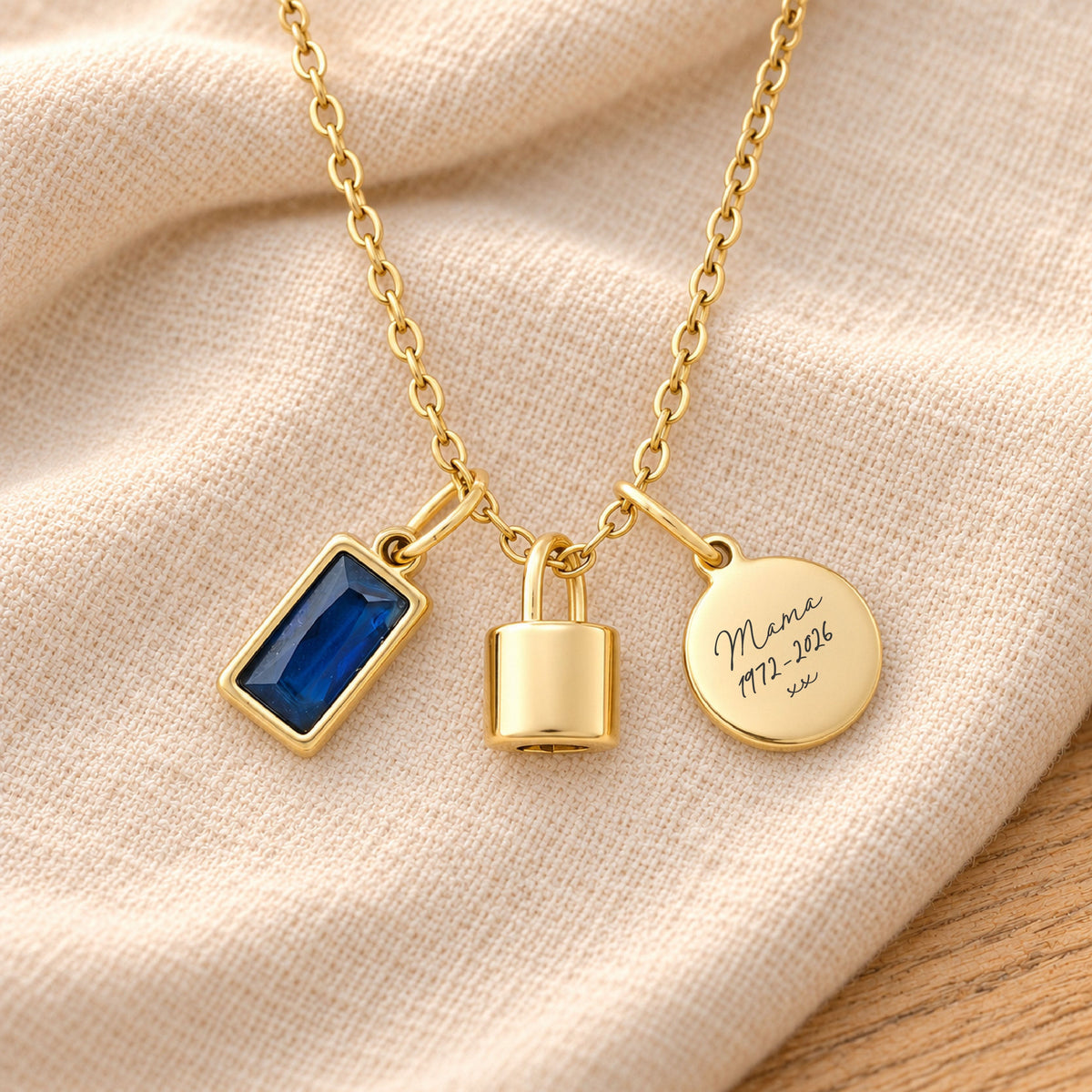 Gold-plated necklace with a rectangular deep blue birthstone charm, a polished lock-shaped urn pendant and a blank round tag, resting on beige linen over rustic wood.