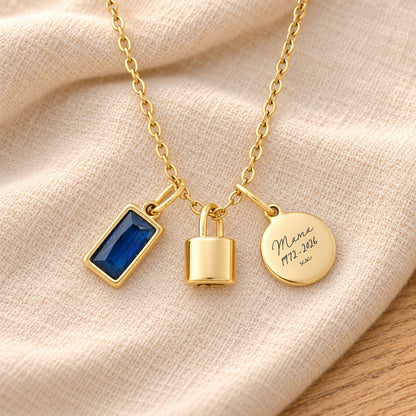 Gold-plated necklace with a rectangular deep blue birthstone charm, a polished lock-shaped urn pendant and a blank round tag, resting on beige linen over rustic wood.