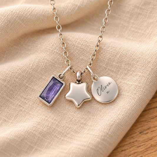 Silver-plated necklace with a rectangular purple birthstone charm, a rounded star urn pendant and a round tag engraved with “Olivia” and a small kiss, resting on beige linen.