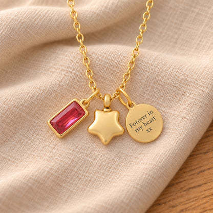 Gold-plated necklace with a rectangular red birthstone charm, a rounded star urn pendant and a round tag engraved with “Forever in my heart xx”, resting on beige linen.