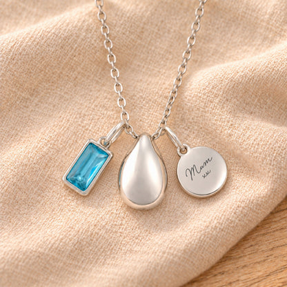 Silver-plated necklace with a rectangular blue birthstone charm, a smooth teardrop urn pendant and a blank round tag, resting on draped beige linen.