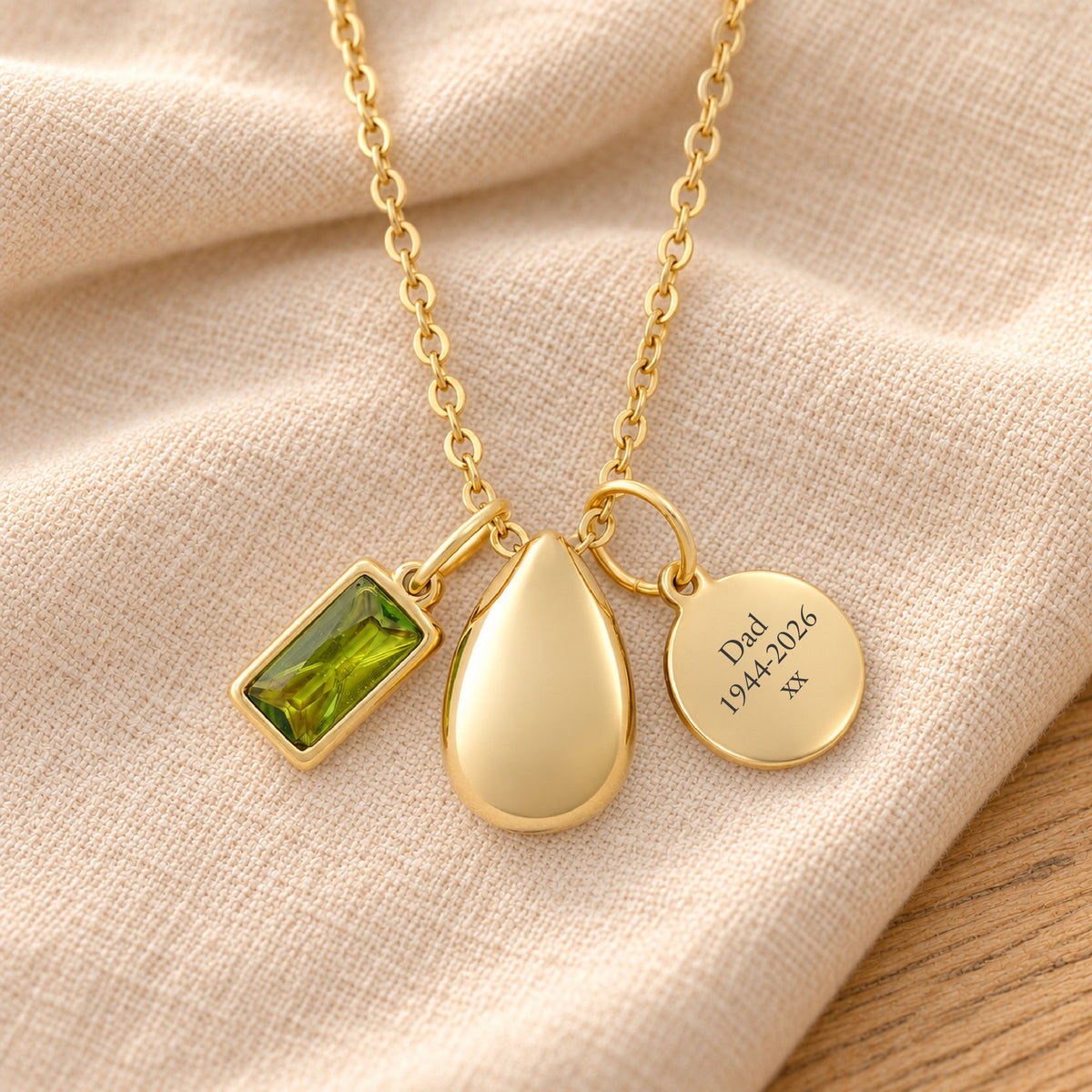 Gold-plated necklace with a rectangular yellow-green birthstone charm, a smooth teardrop urn pendant and a blank round tag, resting on draped beige linen.