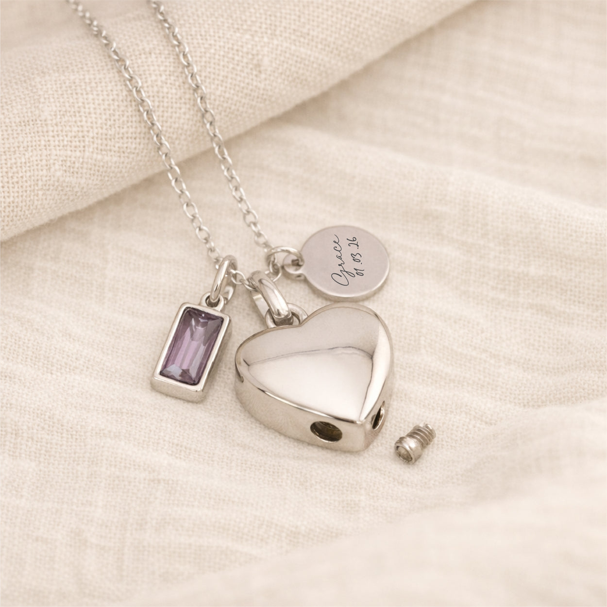 Silver self fill necklace with heart urn pendant, birthstone charm and engraved tag on a linen background