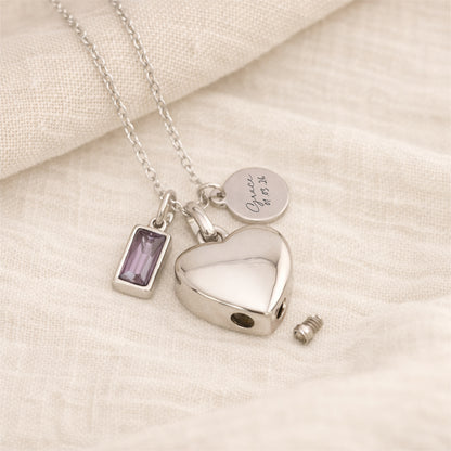 Silver self fill necklace with heart urn pendant, birthstone charm and engraved tag on a linen background