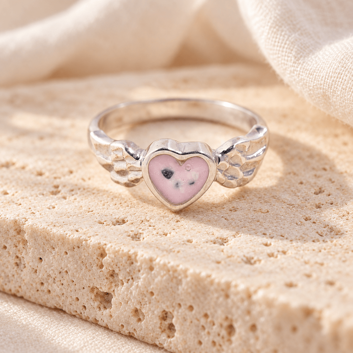 Sterling silver angel wing ashes ring with detailed feather shoulders and a pink resin heart containing visible ashes, resting on cream stone.