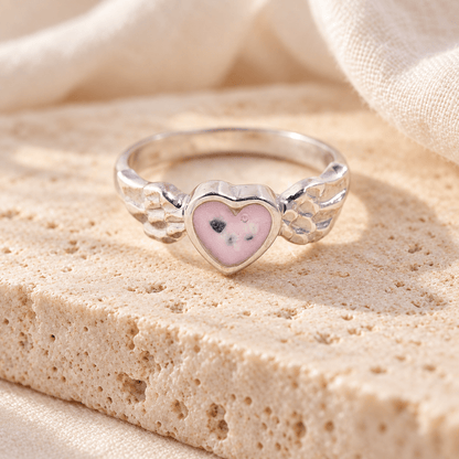 Sterling silver angel wing ashes ring with detailed feather shoulders and a pink resin heart containing visible ashes, resting on cream stone.