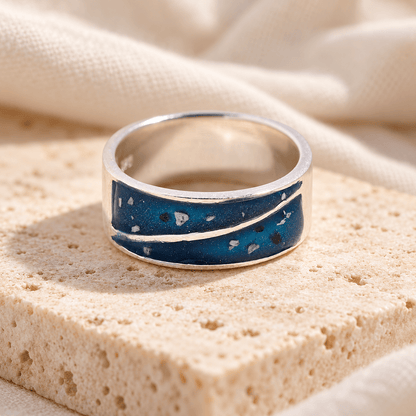 Wide sterling silver duo triangle ashes ring with two deep blue resin sections containing visible ashes, divided by a sweeping silver line, on cream stone.
