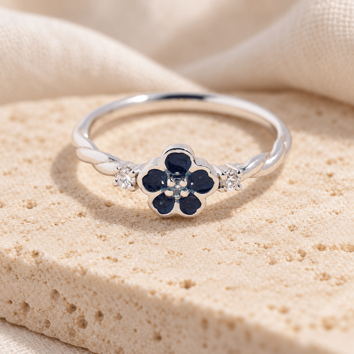 Sterling silver forget me not ashes ring with deep blue resin petals, clear sparkling accents and a twisting band, resting on cream stone.
