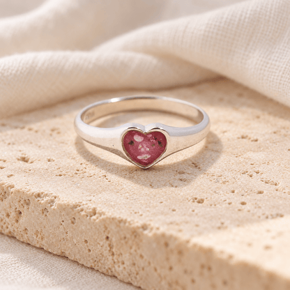Sterling silver heart ashes ring with a smooth tapered band and rose-red resin heart containing visible ashes, resting on cream stone.