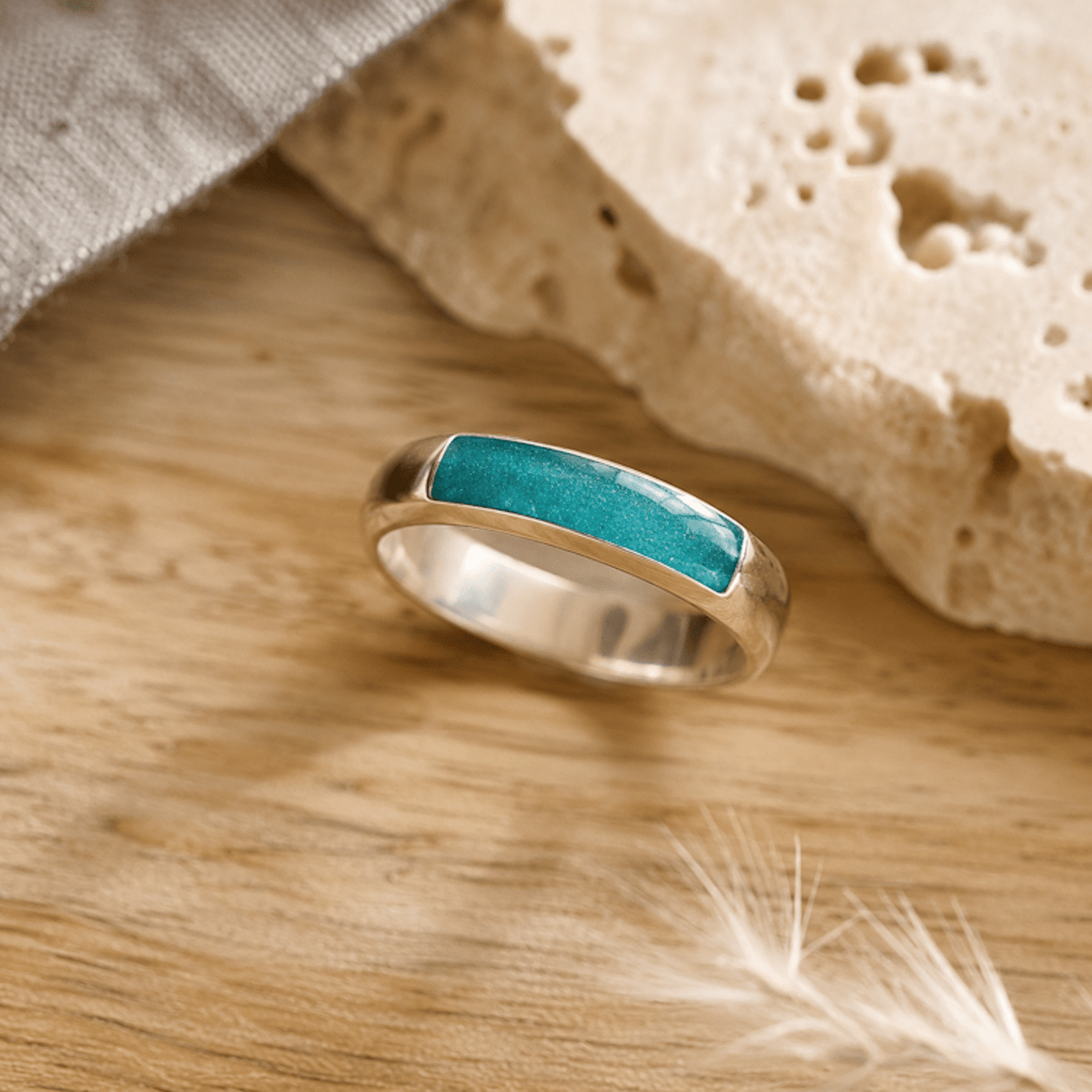 Silver mens ashes ring with teal resin inset for ashes, fur and hair on a wooden surface with a stone and feather.