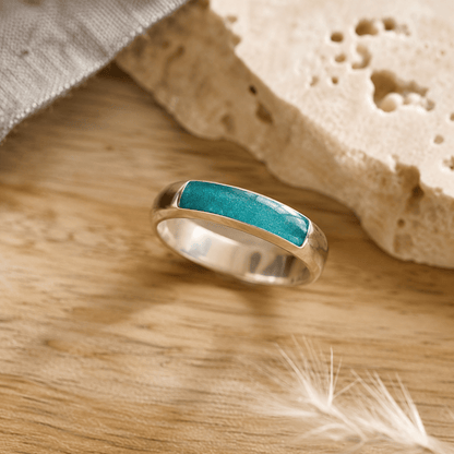 Silver mens ashes ring with teal resin inset for ashes, fur and hair on a wooden surface with a stone and feather.
