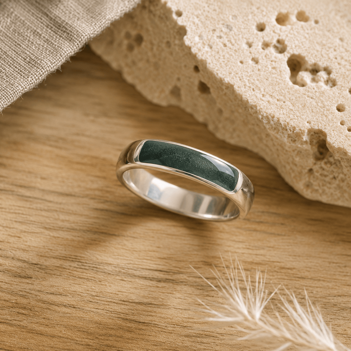 Silver mens ashes ring with evergreen resin inset for ashes, fur and hair on a wooden surface with a stone and feather.