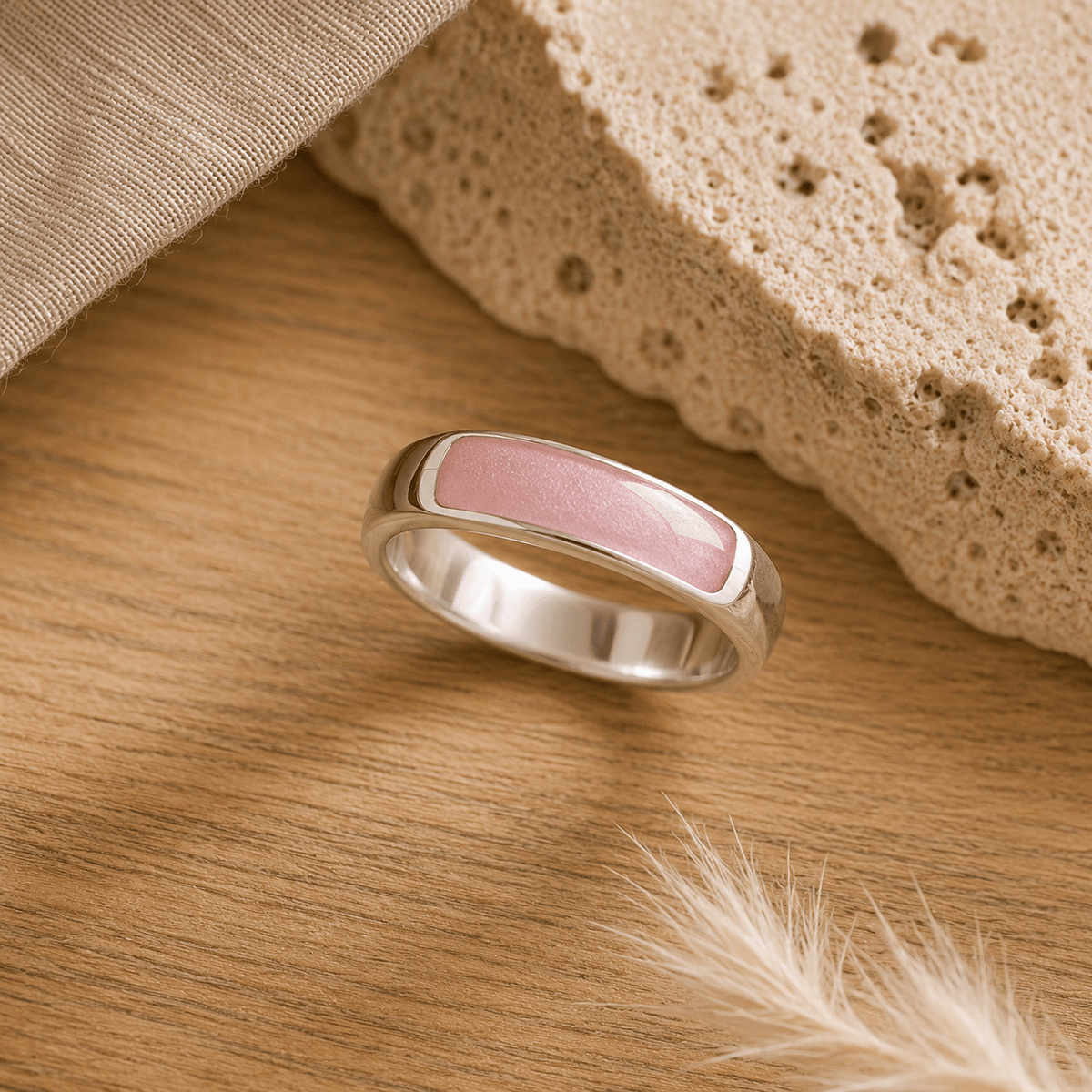 Silver mens ashes ring with blush resin inset for ashes, fur and hair on a wooden surface with a stone and feather.