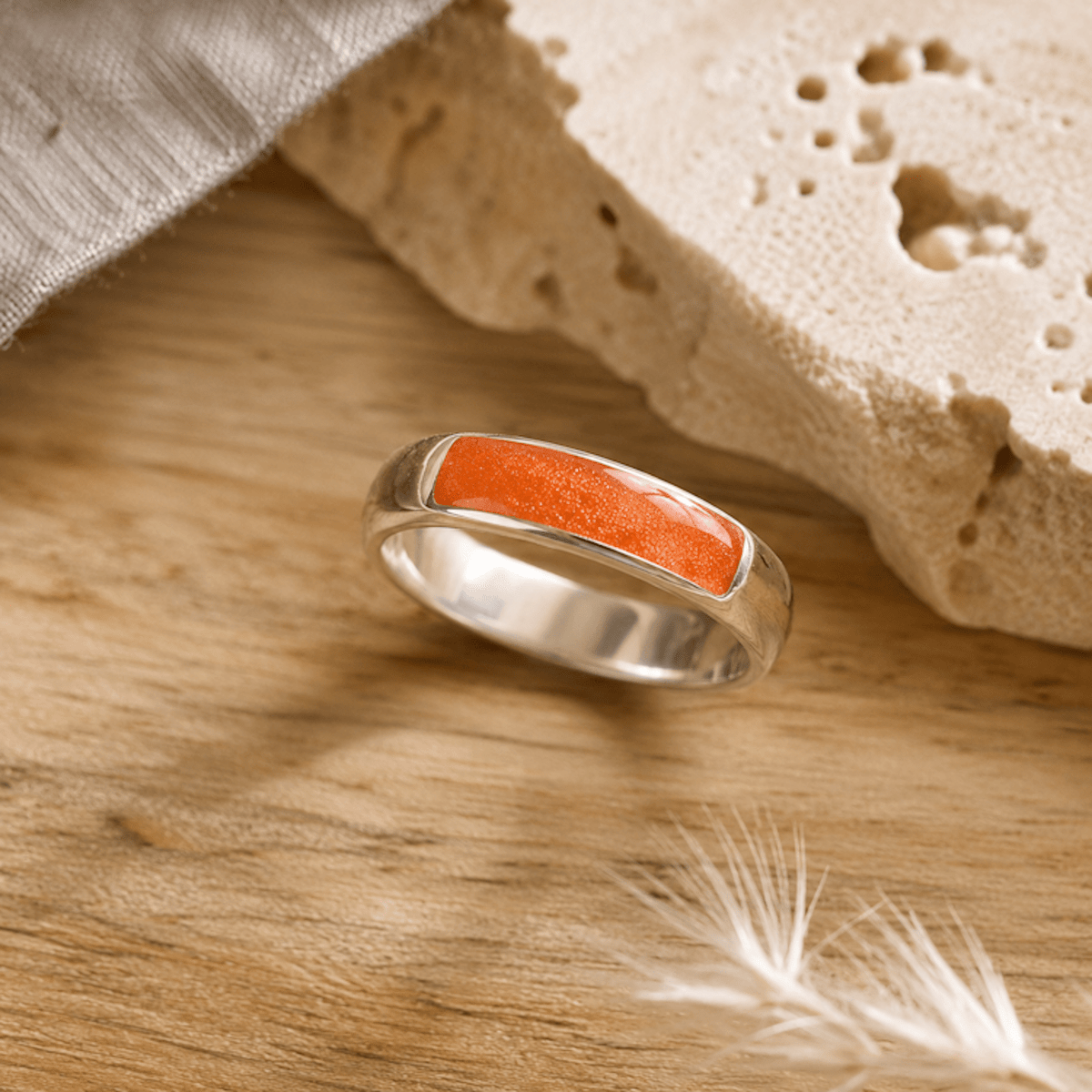 Silver mens ashes ring with orange resin inset for ashes, fur and hair on a wooden surface with a stone and feather.