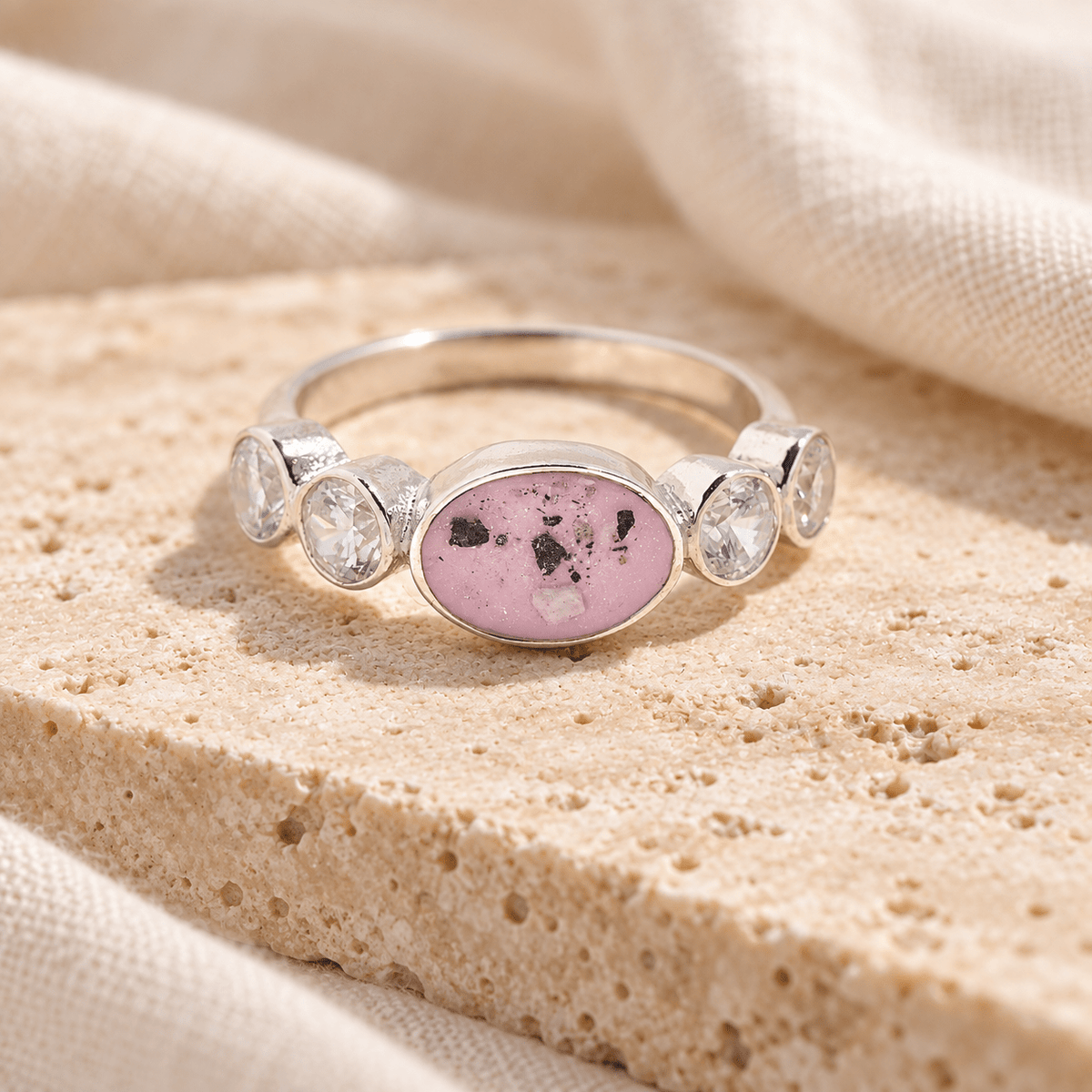 Sterling silver ashes ring with a horizontal pink resin oval containing visible ashes and four clear sparkling accents, resting on cream stone.