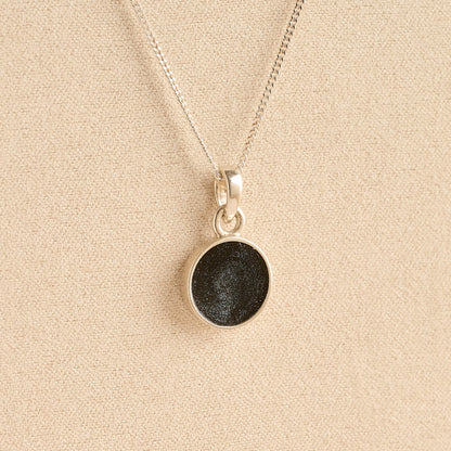 Silver Cremation ashes necklace with a round pendant and black resin filled with ashes or fur, displayed on a soft beige background – suitable for pet or human memorials.