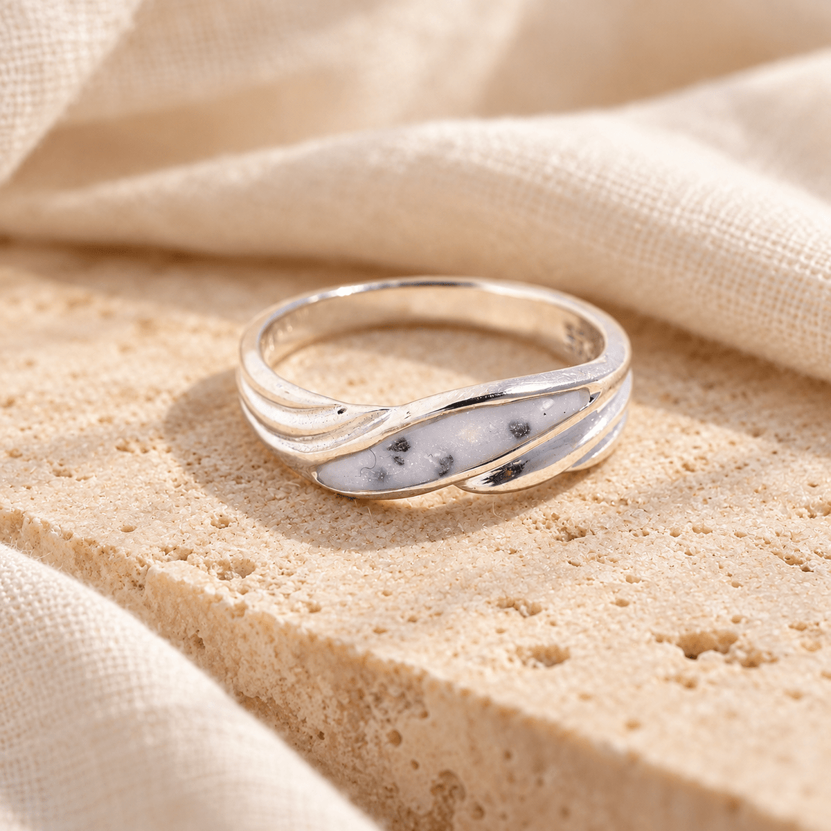 Sterling silver wave ashes ring with flowing polished curves around a pearly white resin setting containing visible ashes, resting on cream stone.