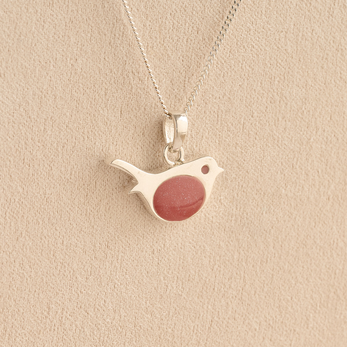 Sterling silver robin pendant with a shimmering reddish-pink resin chest, hanging from a silver chain against a textured beige background.