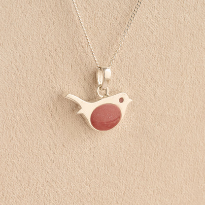 Sterling silver robin pendant with a shimmering reddish-pink resin chest, hanging from a silver chain against a textured beige background.