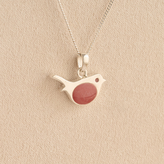 Sterling silver robin pendant with a shimmering reddish-pink resin chest, hanging from a silver chain against a textured beige background.