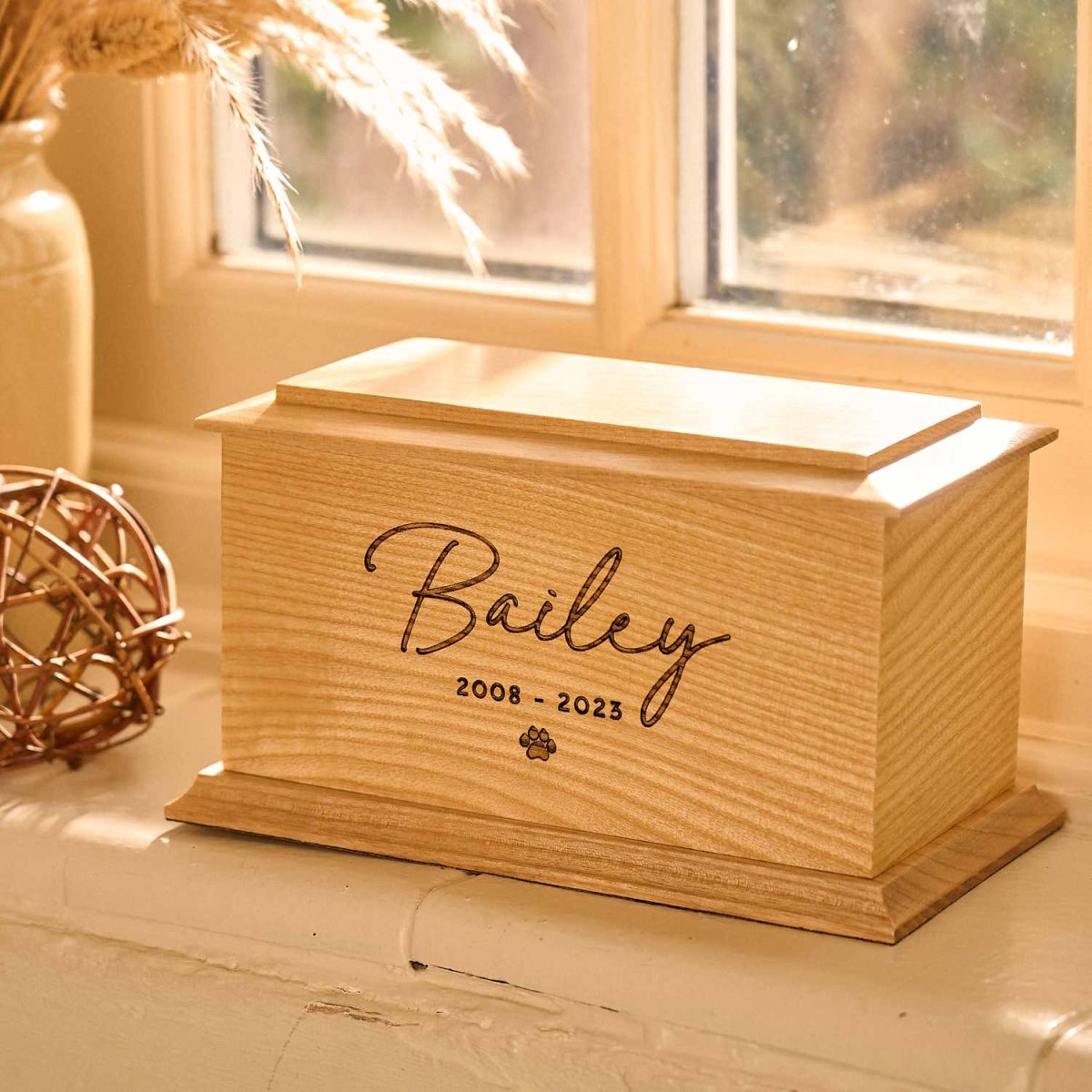 Personalised wooden pet cremation urn with paw print engraving sitting on a windowsill. Suitable as a dog urn or cat urn for pet ashes.