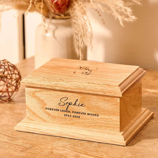 A personalised cremation urn sits on a wooden table. The urn is rectangular and is engraved on the front face with the words "Sophie - Forever Loved, Forever Missed 2015-2023". On the top face is a large capital letter S which has a floral design. It is an urn for human ashes.