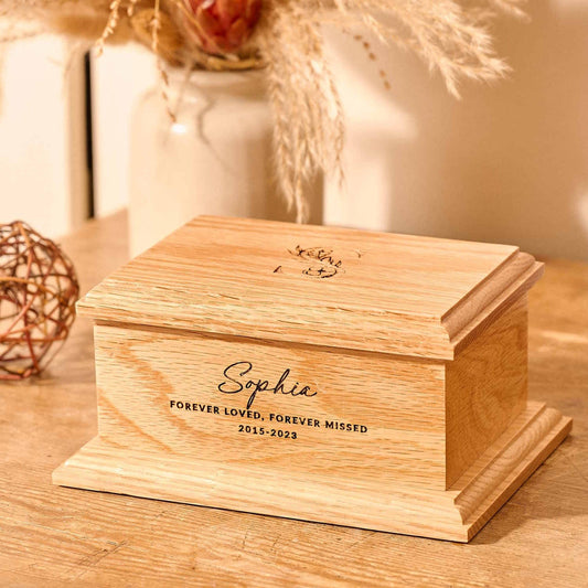 A personalised cremation urn sits on a wooden table. The urn is rectangular and is engraved on the front face with the words "Sophie - Forever Loved, Forever Missed 2015-2023". On the top face is a large capital letter S which has a floral design. It is an urn for human ashes.