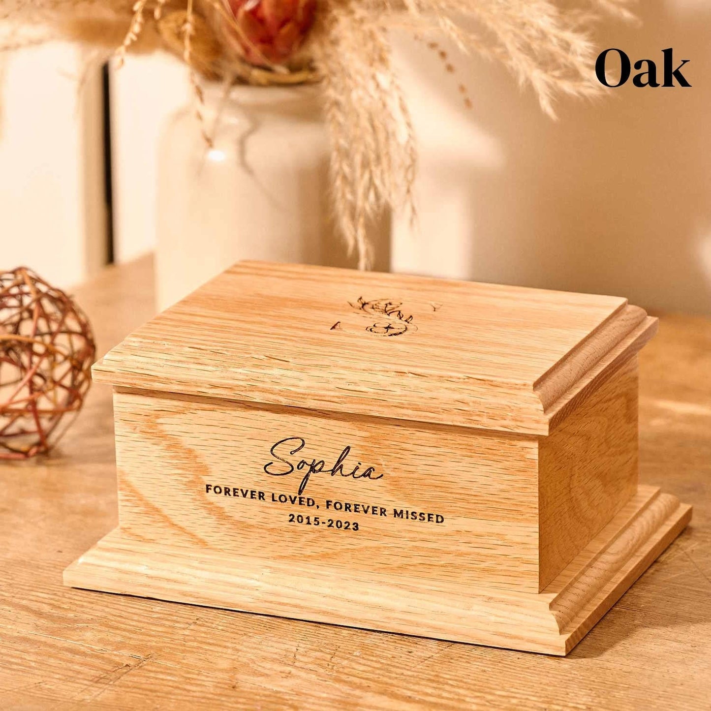 An personalised children's cremation urn sits on a wooden table. The urn is rectangular and is engraved on the front face with the words "Sophie - Forever Loved, Forever Missed 2015-2023". On the top face is a large capital letter S which has a floral design. It is an urn for human ashes.